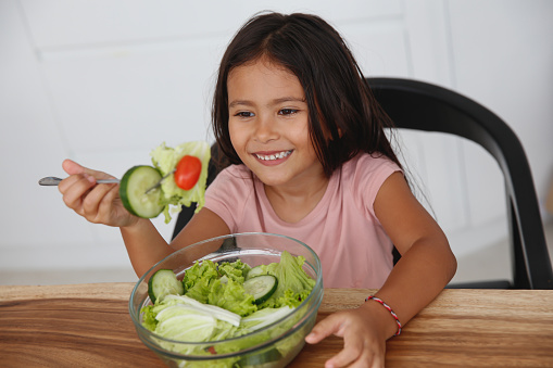 Close-up shot of cheerful multiracial little girl eating healthy green salad at home.