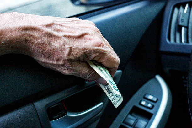 A senior adult man car driver's hand is holding an American twenty dollar bill as he waits in an off-camera fast food restaurant's drive-thru lane to pay for an on the go take out meal.