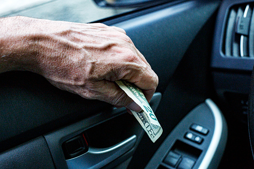 A senior adult man car driver's hand is holding an American twenty dollar bill as he waits in an off-camera fast food restaurant's drive-thru lane to pay for an on the go take out meal.