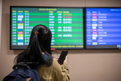 A back view shot of an Asian female passenger checking her boarding time on a monitor screen in the airport.