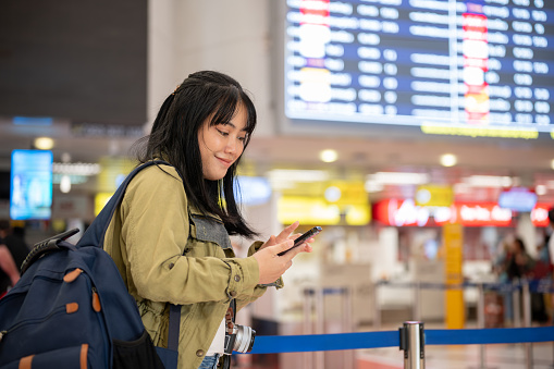 A happy young Asian female traveler or backpacker is using her phone while walking in the airport, checking her flight information.