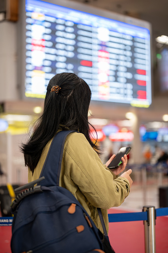 A back view shot of an Asian female traveler or backpacker checking her boarding time on a monitor screen in the airport.