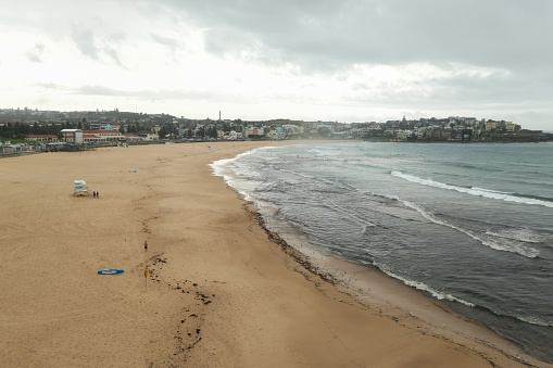 Thunderstorms had passed through the day and night before, and just before this image.  One man of Caucasian appearance is running along the beach; while two men of African appearance and one woman of Caucasian appearance are next to the lifesaver post.  Blue lifesaver surfboards are placed along the beach. Seaweed and pollution have been deposited at the water's edge.  This image was taken on a rainy and overcast afternoon on 20 February 2024.