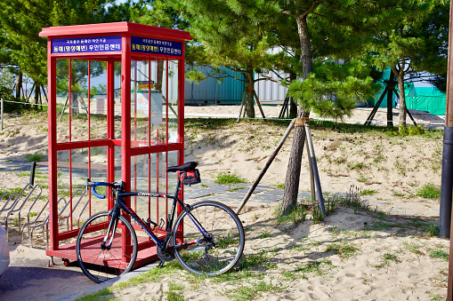 Donghae City, South Korea - July 29th, 2019: A unique red phone booth serves as the Mangsang Beach Certification Center, where cyclists stamp their passports to track adventures along Korea's cross-country routes, set against sandy grounds and pine trees.