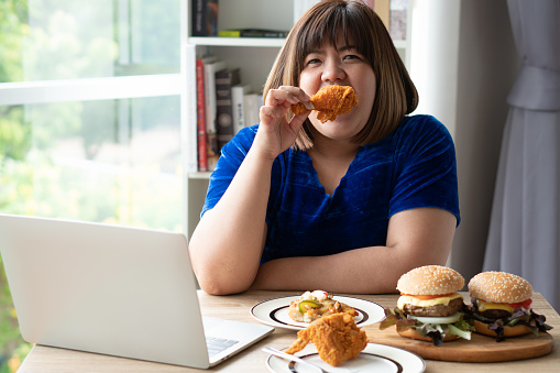 Hungry overweight woman holding Fried Chicken, hamburger on a wooden plate and Pizza on table, During work from home, gain weight problem. Concept of binge eating disorder BED.