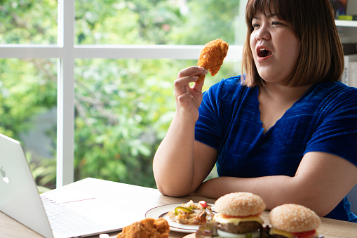 Hungry overweight woman holding Fried Chicken, hamburger on a wooden plate and Pizza on table, During work from home, gain weight problem. Concept of binge eating disorder BED.