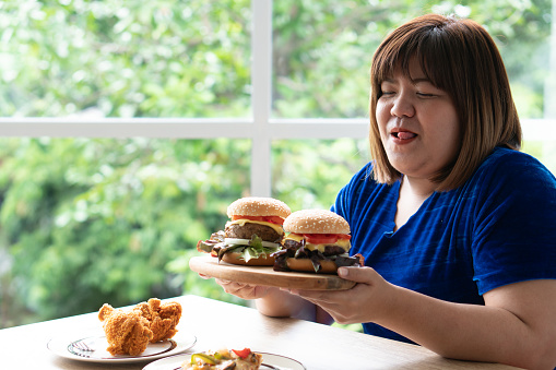 Hungry overweight woman holding hamburger on a wooden plate, Fried Chicken and Pizza on table, During work from home, gain weight problem. Concept of binge eating disorder BED.