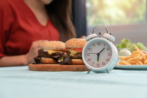 Selective focus of clock, Young woman ready to eating a hamburger, French fries, for Breakfast. Concept of binge eating disorder (BED) and Relaxing with Eating junk food and unhealthy foods.