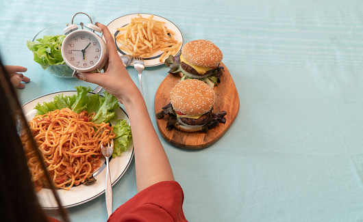 Young woman holding clock and ready to eating a hamburger, French fries, and Spaghetti for Breakfast. Concept of binge eating disorder BED and Relaxing with Eating junk food and unhealthy foods.