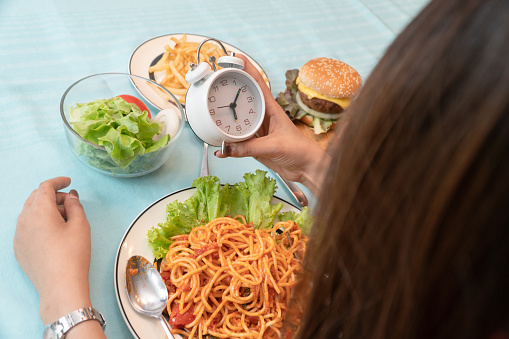 Young woman holding clock and ready to eating a hamburger, French fries, and Spaghetti for Breakfast. Concept of binge eating disorder (BED) and Relaxing with Eating junk food and unhealthy foods.