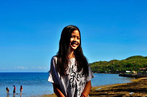 A portrait of a young teenage girl at the foreground against a scenic background of the coastline where beachgoers bathe in the sea and collect sea shells on the shore.