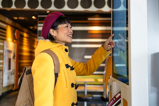 Woman in trendy coat checking menu and ordering meal on self service interactive display inside fast food restaurant.