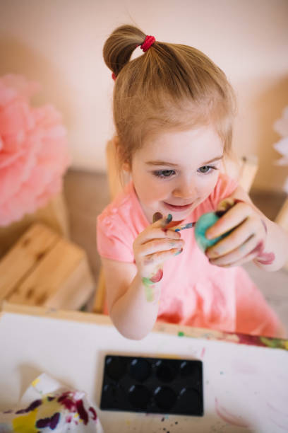 Cute happy 3 years old girl wearing pink dress painting Easter eggs in bright colourful room