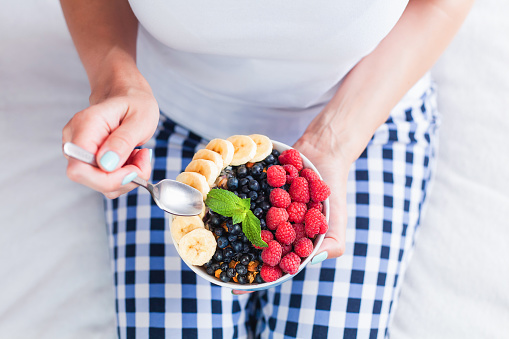 The girl eats a healthy breakfast of muesli with fresh fruits and berries while sitting on the bed at home. Healthy food concept. ?lose-up. Top view.