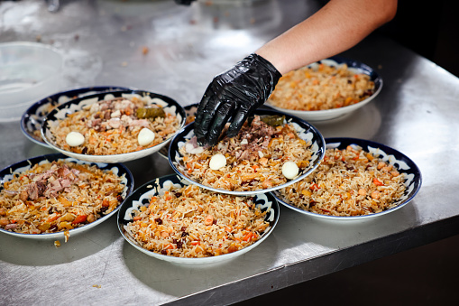 A person wearing black gloves is seen carefully scooping and portioning out food into bowls. The focus is on the precise and meticulous actions involved in cooking a dish, likely the national dish of Uzbekistan, pilaf.