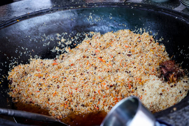A pan filled with food is placed on top of a stove, where it is being cooked. The dish being prepared is Pilaf, the national dish of Uzbekistan, known for its flavorful combination of rice, meat, vegetables, and spices.