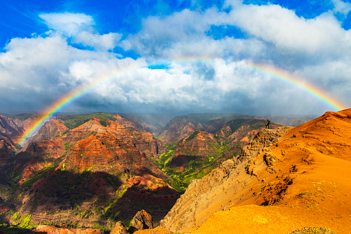 Young man overlooking rainbow over majestic and colorful mountains and valleys in Hawaii. Photographed in Waimea Canyon, Kauai, Hawaii.