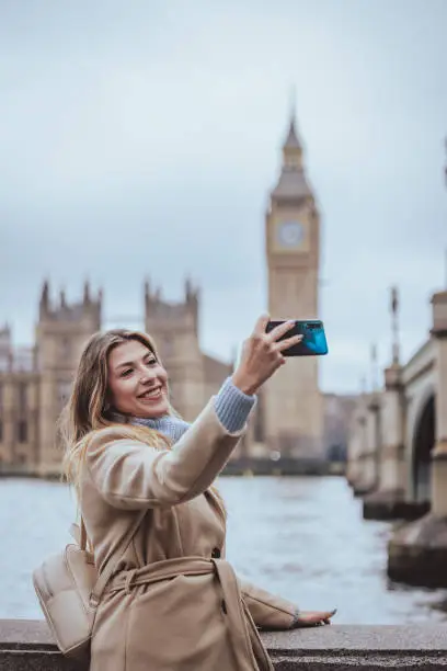 Candid waist up portrait of a young cheerful blond woman in her 30s taking selfie with her smart mobile phone device while smiling at the screen in down town city of London, England. Selective focus on the model with plenty of copy space on the background and sky, which is defocused Westminster Bridge, Parliament building and Big Ben clock tower, city of London, Great Britain. Photo created during cold season outdoors and the model is with warm casual clothes in cream and light blue colours - creative stock photo