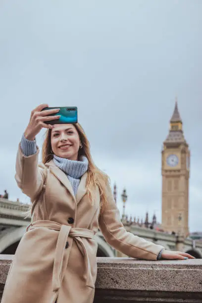 Candid waist up portrait of a young cheerful blond woman in her 30s taking selfie with her smart mobile phone device while smiling at the screen in down town city of London, England. Selective focus on the model with plenty of copy space on the background and sky, which is defocused Westminster Bridge, Parliament building and Big Ben clock tower, city of London, Great Britain. Photo created during cold season outdoors and the model is with warm casual clothes in cream and light blue colours - creative stock photo