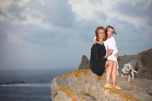 A tender little girl embraces her mother. They stand on a towering boulder at Capo Testa, overlooking the sea. The sky looks gloomy, covered with storm clouds.