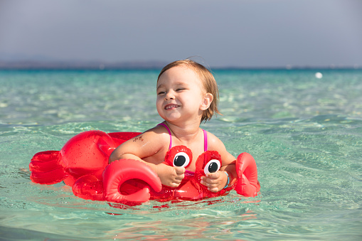 Just a few meters from the center of Santa Teresa Gallura, the transparent and colorful sea will amaze any visitor. Here is the magnificent white-sand Rena Bianca beach. A little girl enjoys swimming with a red life buoy in the shape of a crab. In the background, the white cliffs of Bonifacio and the mountains of Corsica.