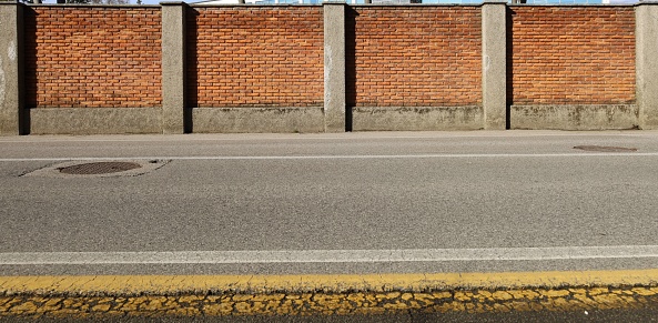 High surrounding brick wall with concrete columns at the road side. Empty large street in front with road line markings. Urban background for copy space.