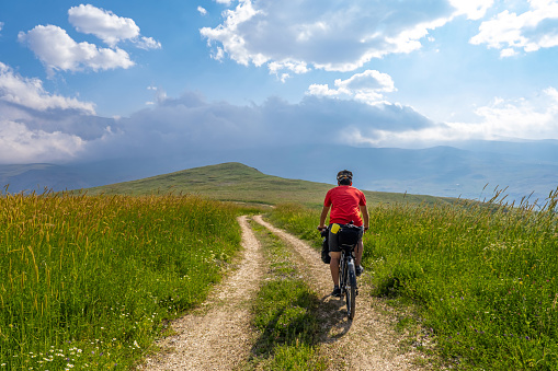 Curvy dirt road going down to the valley among the meadows. Sunset, cloudy weather, mountainous environment. Red-topped cyclist. Nature view photo with high adventure spirit from the back