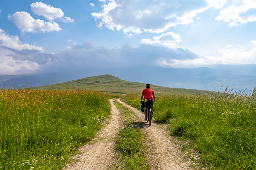 Curvy dirt road going down to the valley among the meadows. Sunset, cloudy weather, mountainous environment. Red-topped cyclist. Nature view photo with high adventure spirit from the back