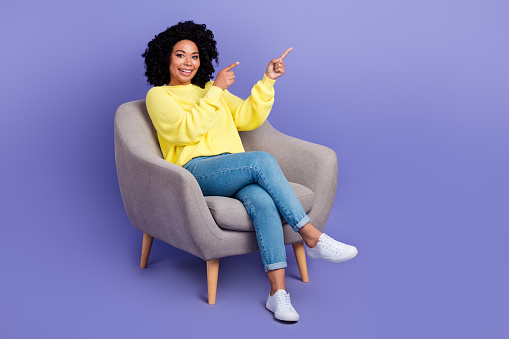 Full size photo of optimistic woman dressed sweater sit on armchair indicating at discount empty space isolated on violet color background.