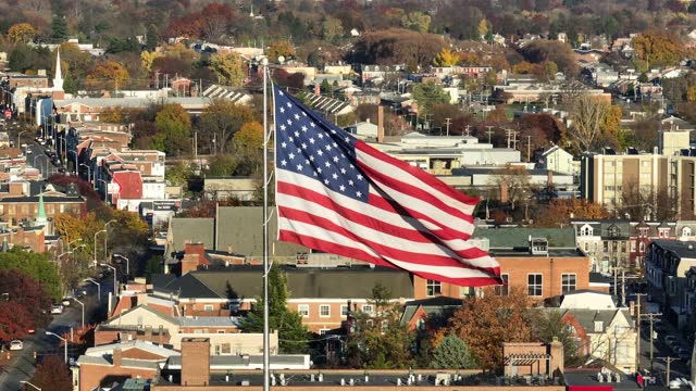 American flag waving over urban cityscape with autumn foliage. Aerial establishing shot of Northeast town with patriotic theme.