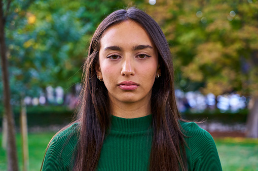 Portrait of a young brunette woman looking at camera with a serious face gesturing disappointment and discomfort while standing outdoors in a park.