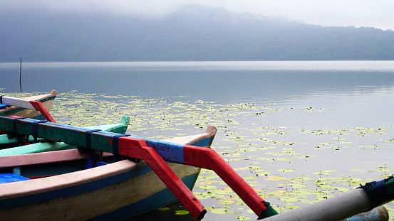 The tranquil view of the lake with calm water and peaceful morning atmosphere. Closeup part of colorful wooden fishing boats floating on lake. Beratan Lake Bedugul, Bali, Indonesia.