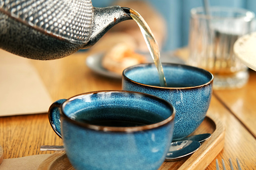 Brewing and Serving Tea. Beautiful Teapot Pouring Green Tea in Blue Ceramic Cup on Wooden Cafe Table. Two Pottery Handmade Mugs Early in the Morning. Trendy Authentic Porcelain Set. Breakfast Concept. Tea Time. Trend of stylish Dishware, Tableware. High tea