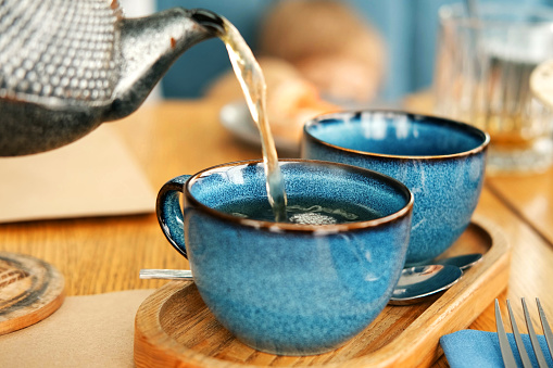 Brewing and Serving Tea. Beautiful Teapot Pouring Green Tea in Blue Ceramic Cup on Wooden Cafe Table. Two Pottery Handmade Mugs Early in the Morning. Trendy Authentic Porcelain Set. Breakfast Concept. Tea Time. Trend of stylish Dishware, Tableware. High tea