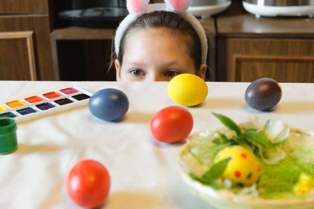 Cute little girl wearing bunny ears playing egg hunt on Easter, hides under the table and picks up the egg. Adorable children celebrate Easter at home, Close up. Happy Easter. Eggs Hunt. 4K
