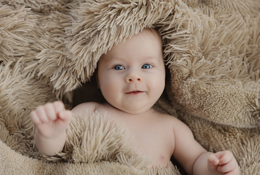 Cute happy smiling baby girl lying on beige blanket, looking at camera. Top view on little infant on bed covered with plaid, rug. Kid looks excited, amused expression, close-up. Child three months old