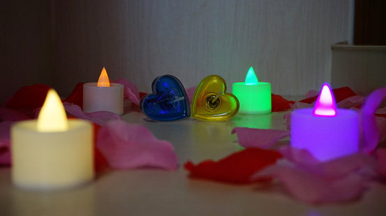 There are two heart shaped candels among four different colored candels. The are red and pink color rose petals in the foreground and background of the candles.