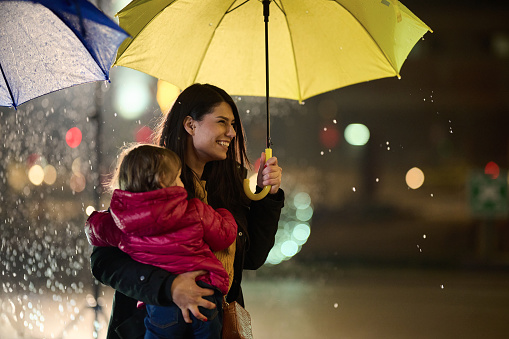 A mother tenderly carries her little daughter while shielding her with an umbrella on a rainy night, embodying the protective love and warmth of maternal care amidst the urban cityscape.