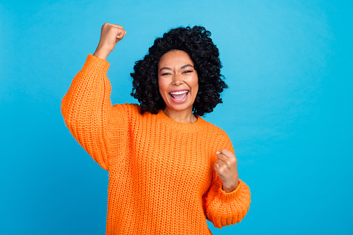 Photo portrait of pretty young girl raise fist celebrate achieve dressed stylish knitted orange outfit isolated on blue color background.