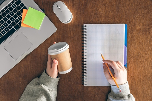 Woman with a cup of coffee writes in a notepad on a wooden table, top view. Minimal work space, creative flat lay photo of workspace desk.