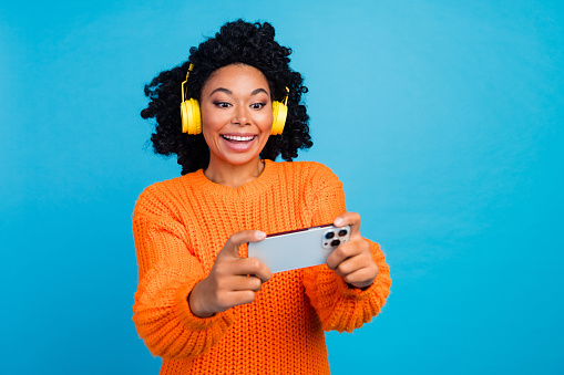 Photo portrait of pretty young girl play mobile game headphones dressed stylish knitted orange outfit isolated on blue color background.