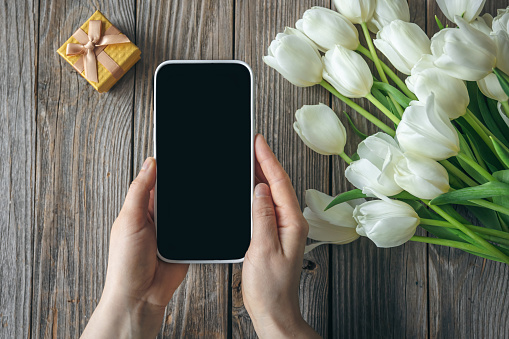A smartphone in female hands, a yellow gift box and a bouquet of white tulip flowers on a wooden background, top view.