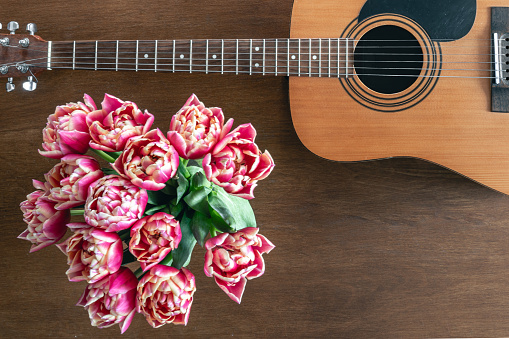 Bouquet of pink tulips and acoustic guitar on a wooden background, top view. Spring flowers and guitar on the table. Copy space.