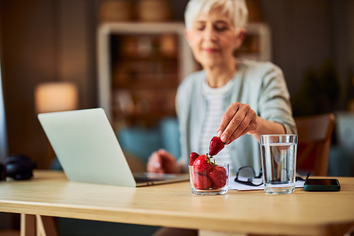 A senior adult woman having a strawberry as a snack while sitting at a desk in front of a laptop and taking a break from work.