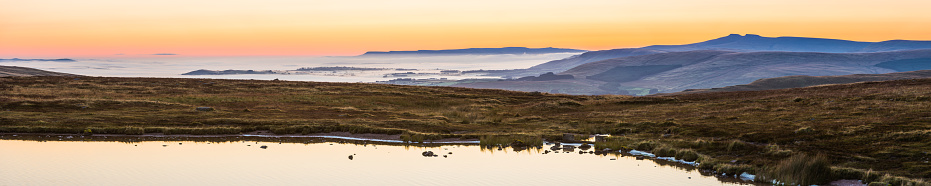 Tranquil wilderness lake shore as the first light of dawn brightens over the mist filled valleys and mountain ridges on the horizon of the Brecon Beacons National Park, Wales.