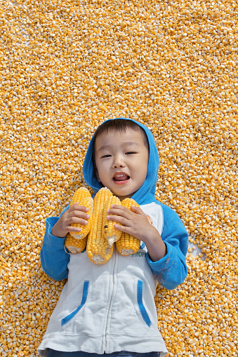 A little Asian boy, lying flat on his back in a pile of corn kernels outside during the day, looking at the camera with a smile.