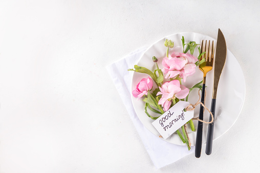 Spring holiday table setting with tender cute rose pink flowers, cutlery - plate, knife, fork on white background with tag Good morning, top view copy space