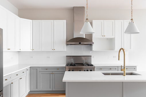 A kitchen detail with grey and white cabinets, gold faucet, gold and white pendant lights hanging above the island, and stainless steel appliances.