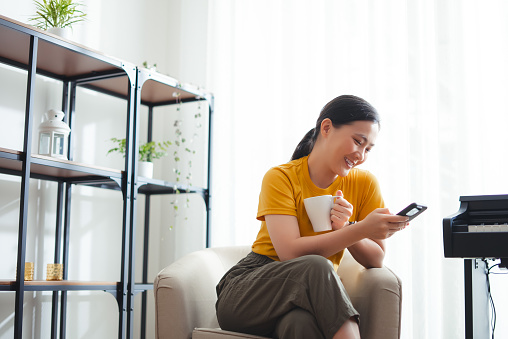 Asian woman sitting on armchair with warm coffee and holding smart phone at home. Happy woman using smart phone for searching shopping online or surfing social media.