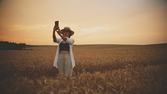 Woman Snaps A Selfie Using Her Mobile Phone In A Wide Wheat Field During Sunset